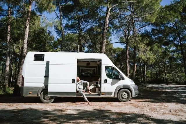 person relaxing at the open door of a white campervan parked in a forest