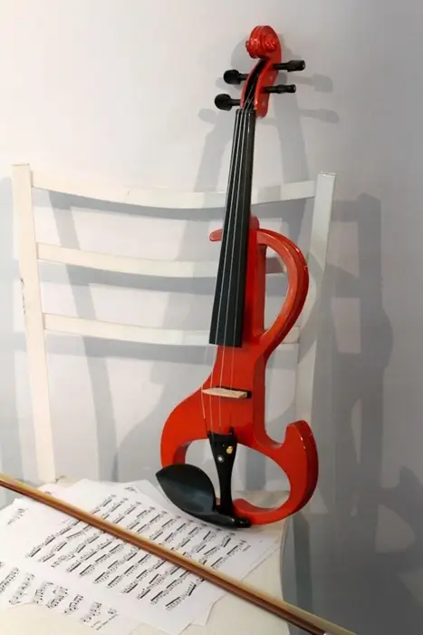 A red electric violin leaning against a white chair with sheet music and a bow on the floor