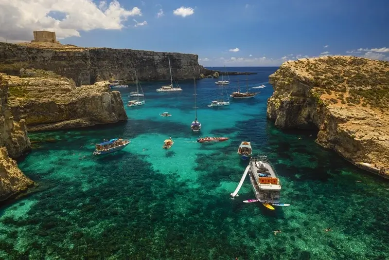 Several boats anchored in a sheltered cove with vivid green-blue water framed by dramatic limestone cliffs and a historic watchtower on Comino island