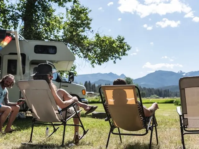 group of friends relaxing on camping chairs beside an RV with mountain views in the background
