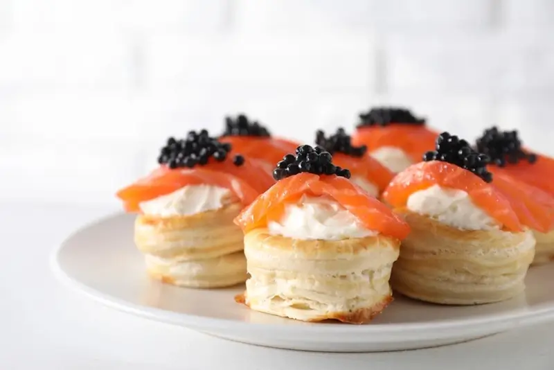 French puff pastry bites topped with smoked salmon, cream cheese, and black caviar on a white plate