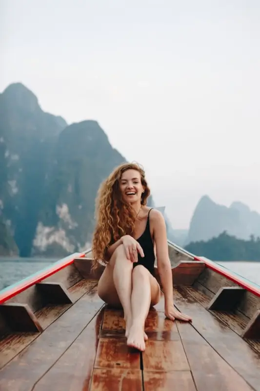 Happy female traveler sitting on a wooden boat surrounded by misty mountains and calm water