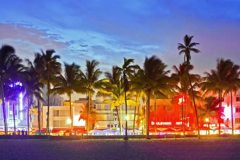 Colorful Art Deco hotels lit up at night along Ocean Drive in Miami Beach with palm trees and a vibrant sunset sky