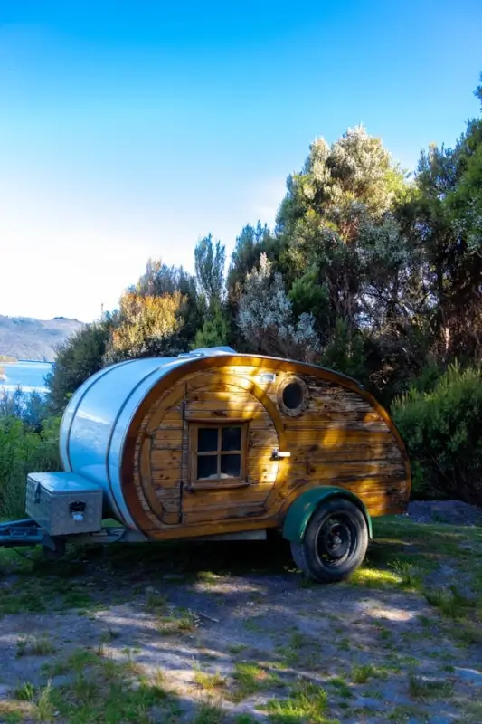 rustic wooden teardrop caravan parked among native Tasmanian shrubs with a glimpse of blue water in the background