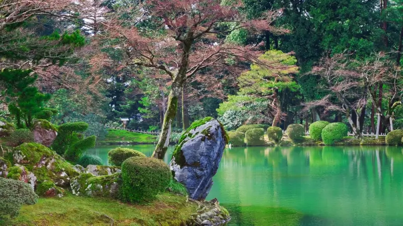 Kasumiga-ike Pond at Kenrokuen Park in Kanazawa, Japan, surrounded by moss-covered rocks, manicured shrubs, and autumn trees reflecting on the still green water