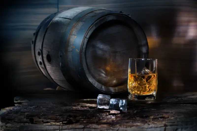 Crystal glass of amber Scottish whisky with ice cubes beside a weathered oak aging barrel