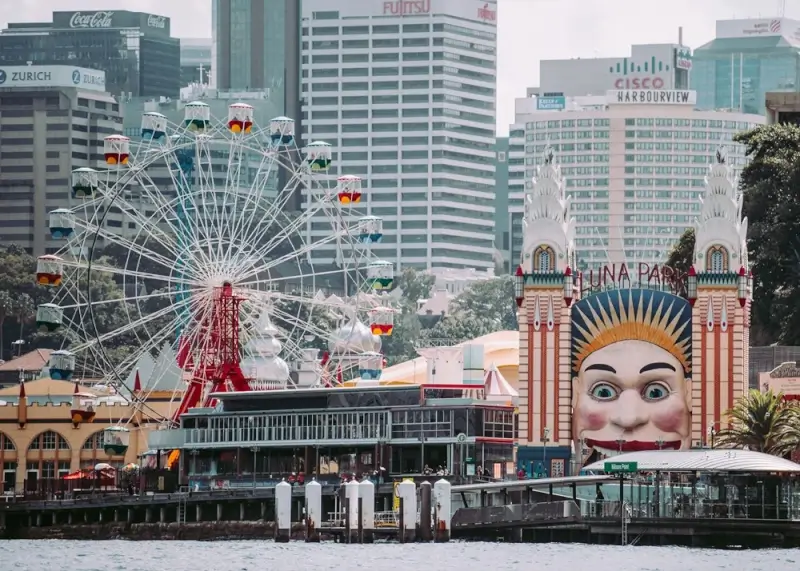 Iconic Luna Park Sydney amusement park seen from the harbour, featuring its famous smiling face entrance, a colorful Ferris wheel, and the city's skyscraper skyline in the background.