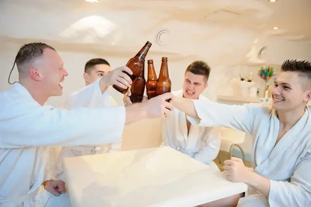 Group of men in white robes toasting with beer bottles during a spa session together