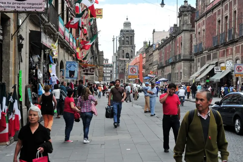 A busy pedestrian street in Mexico City with locals walking past street vendors and Mexican flags, with a historic cathedral visible in the background
