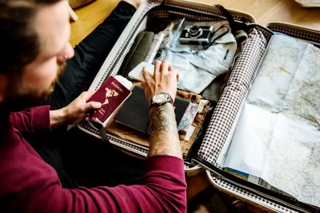 A man holding a passport while packing a suitcase with travel essentials including a camera, maps, and clothes.