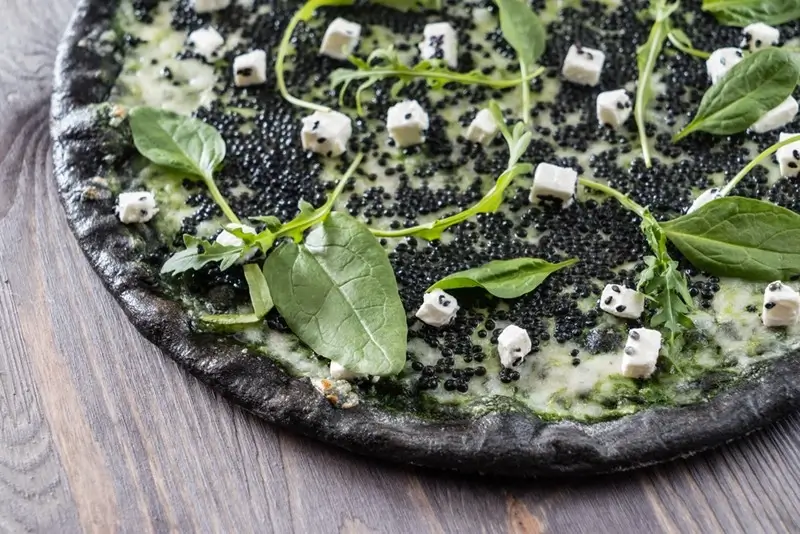 Black squid ink pizza topped with black caviar, feta cheese, and fresh greens on a wooden surface