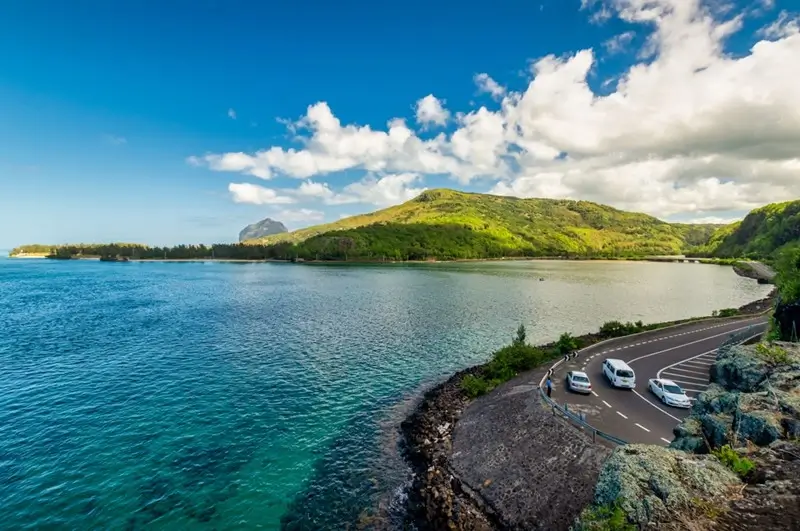 Cars driving along a scenic coastal road in Mauritius with turquoise lagoon and Le Morne Brabant mountain in the distance