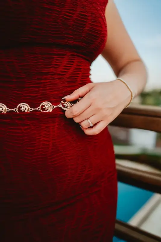 Close-up of a woman in a textured red dress with a gold chain belt, wearing a ring and bracelet, standing on a balcony