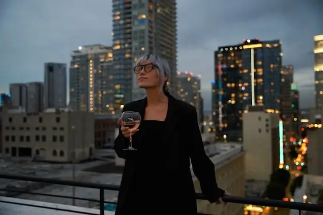 A solo woman enjoying a drink on a rooftop bar overlooking a lit-up city skyline in the evening