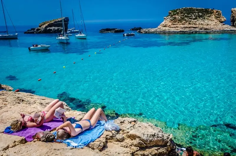 Two women sunbathing on rocky shores beside crystal-clear turquoise water with sailboats and a small rocky islet in the distance