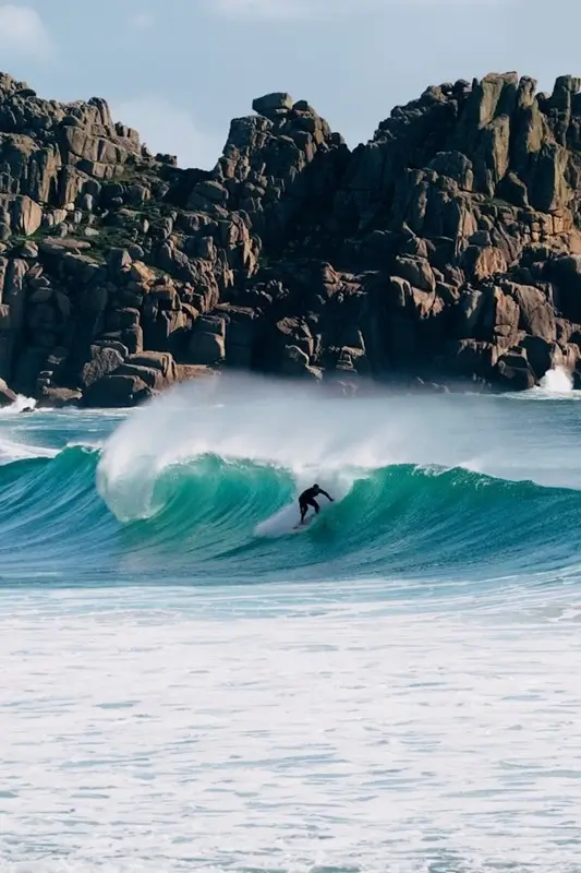 A surfer riding a large turquoise wave along the rugged Cornwall coastline with dramatic granite rock formations in the background