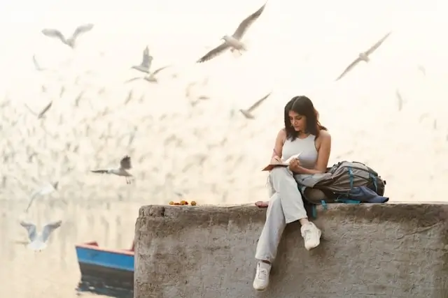 A woman sitting outdoors with a backpack, writing in a journal surrounded by seagulls in flight near a calm waterway