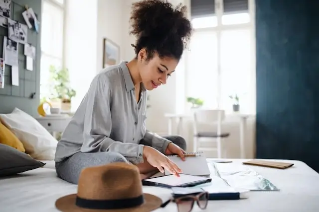 Happy female traveler organizing her trip plans on a bed with a notebook, tablet, sunglasses, and a hat