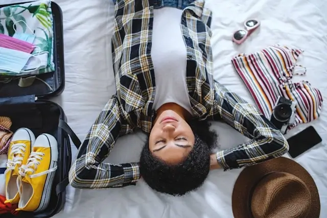 Overwhelmed female traveler lying on a bed surrounded by an open suitcase, shoes, sunglasses, and travel essentials while packing