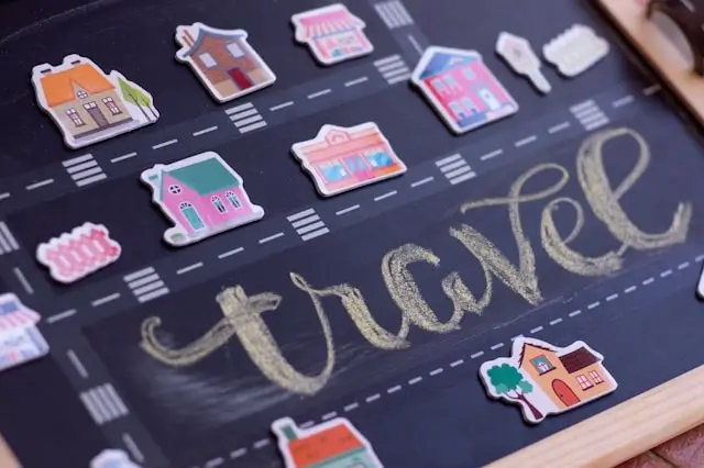 A chalkboard with the word "travel" written in gold lettering surrounded by colorful miniature house stickers along illustrated roads.