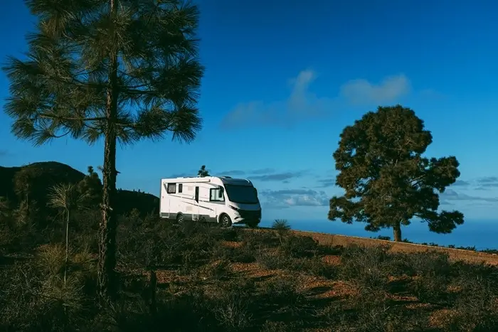 modern white motorhome parked on a hilltop with a person sitting on the roof surrounded by pine trees and blue sky