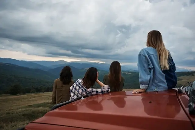 A group of friends sitting on top of a car together, looking out over a scenic mountain landscape during a road trip
