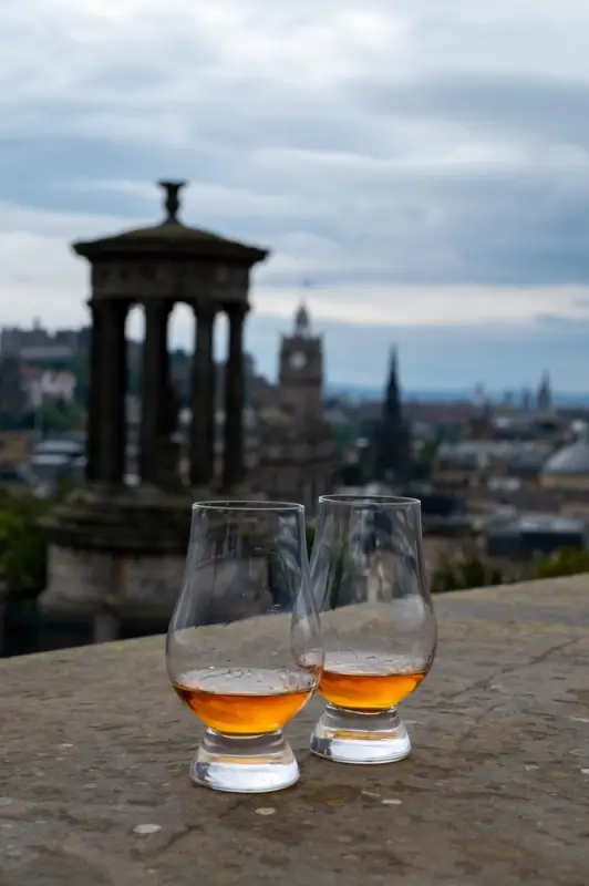 Two Glencairn glasses of Scotch whisky on a stone ledge overlooking the Edinburgh skyline