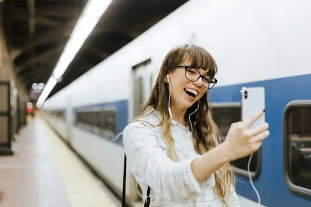 A cheerful woman with glasses and earphones on a video chat on her phone at a train station platform