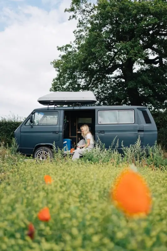 A woman sitting in the open doorway of a camper van parked in a wildflower field, enjoying a quiet moment alone on a solo trip