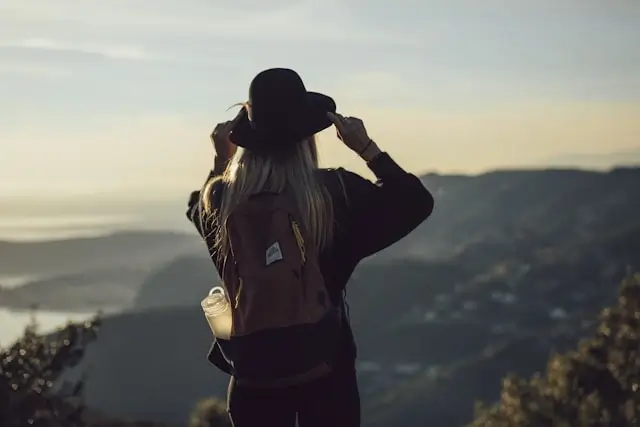 A woman with a backpack standing alone at a mountain viewpoint, looking out over a vast landscape at golden hour
