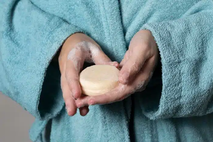 Woman in a bathrobe holding a solid shampoo bar with lathered hands
