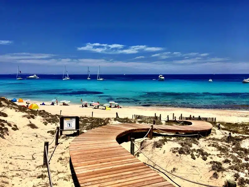 Sunny white sand beach in Formentera with a wooden boardwalk, turquoise water, and sailboats anchored offshore