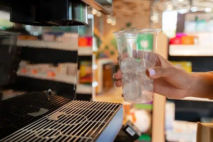 Person holding a clear cup filled with ice next to a commercial slush machine cooling system in a retail setting.