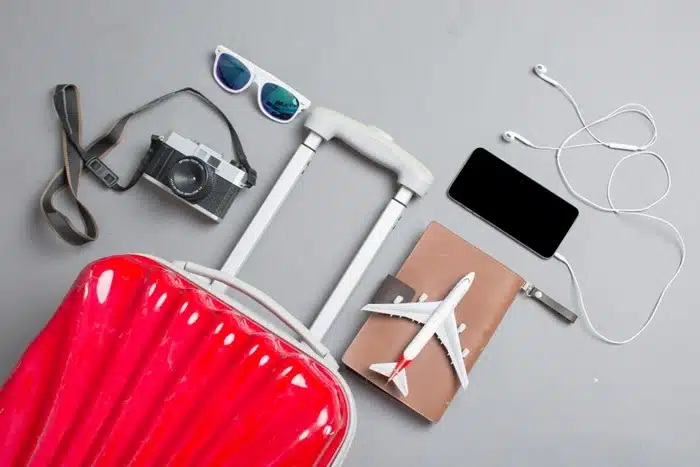 Flat lay of essential travel tools including a red suitcase, camera, passport, phone, and sunglasses
