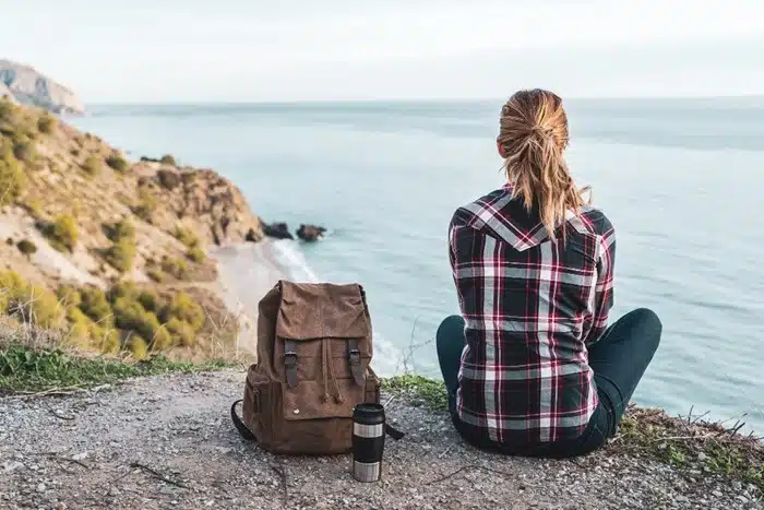 Woman sitting alone on a rocky cliff edge enjoying a secluded ocean view with a travel backpack nearby