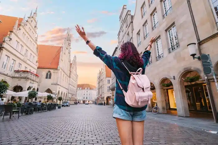 A solo female traveler with a backpack raising her arms freely in a charming European city square at sunset
