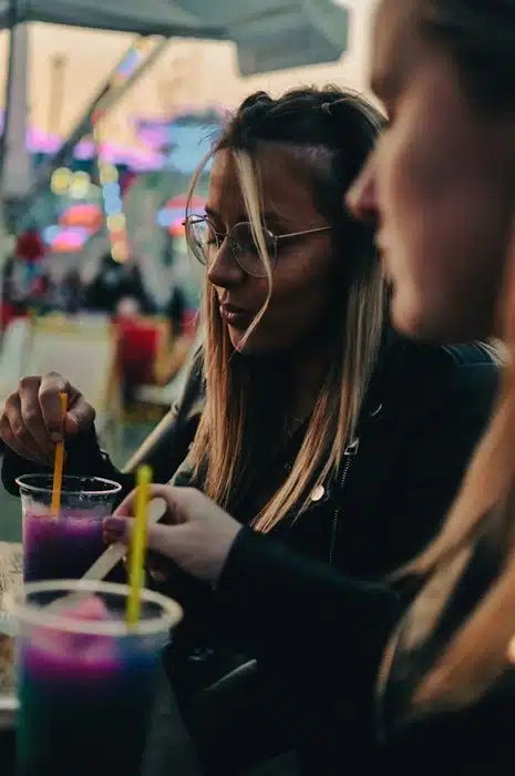Two young women enjoying slushie drinks at an outdoor venue with colorful city lights in the background.