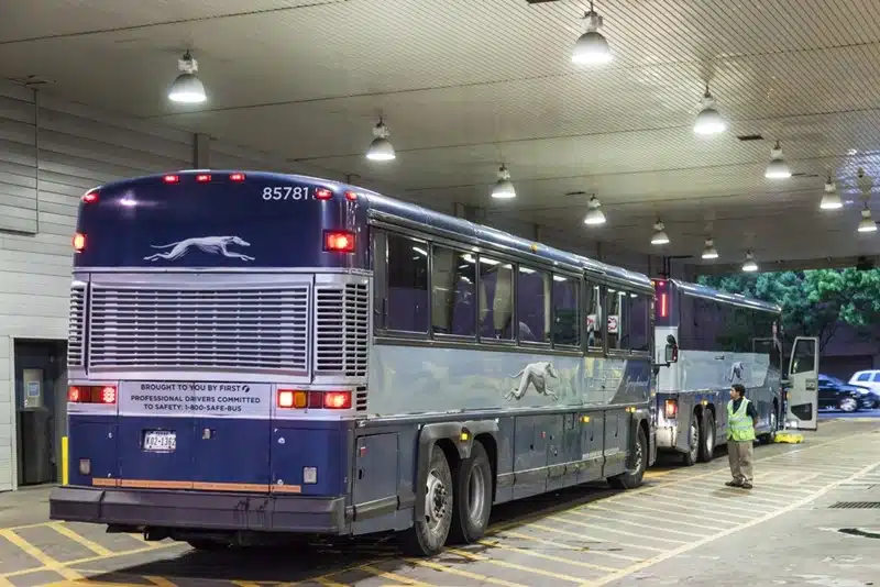 Greyhound buses lined up at a covered terminal at night with a staff member on the platform