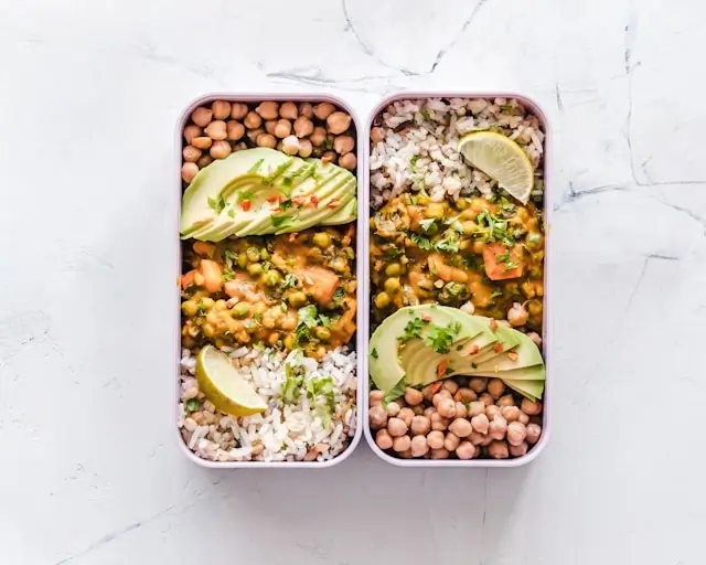 2 meal prep containers filled with rice, chickpeas, avocado, and curry on a marble surface