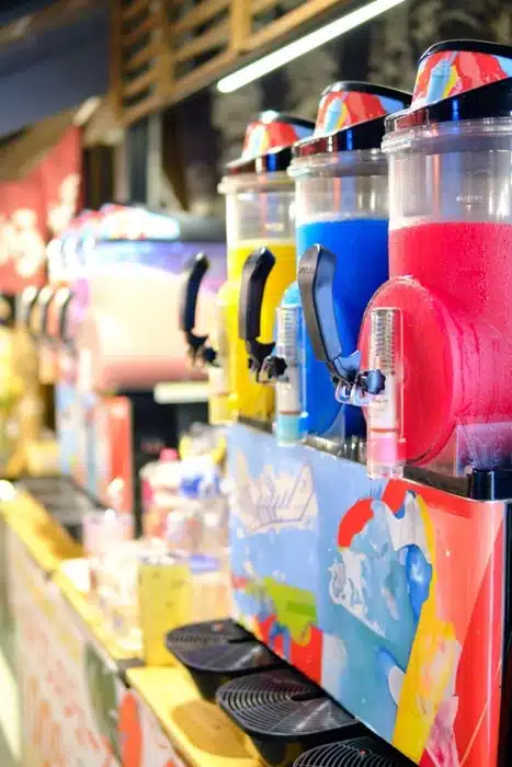A row of colorful commercial machines for slushies lined up at a busy food service counter with multiple flavors on display.