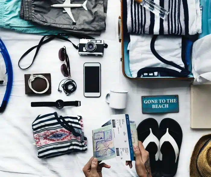 Flat lay of travel packing essentials including passport, camera, phone, flip flops, and open suitcase