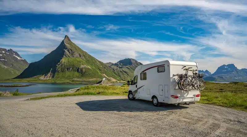 White RV with bikes on the back parked on an open road trip guide route surrounded by mountains and a lake