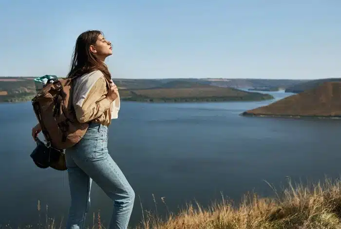 A woman with a hiking backpack standing on a hilltop, enjoying a sweeping view of a river and open landscape on a clear day