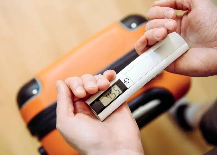 Hands holding a digital luggage scale showing weight in front of an orange suitcase