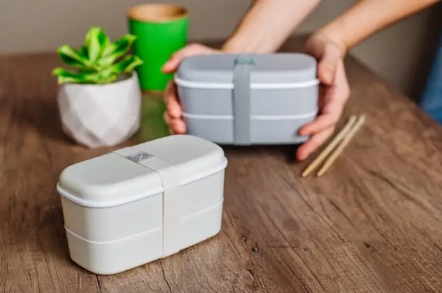 hands holding a stacked lunch box on a wooden table with chopsticks and a small plant in the background