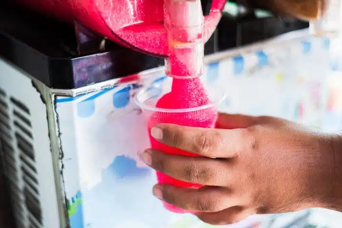 Person filling a plastic cup with a pink slushie drink from a commercial slush machine spigot.