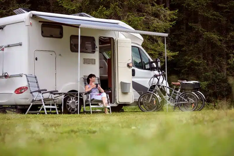 Woman relaxing with a coffee outside her RV parked in a forest clearing with bikes nearby