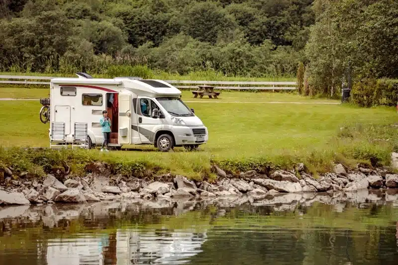 Person standing by an RV parked on a grassy lakeside spot surrounded by trees