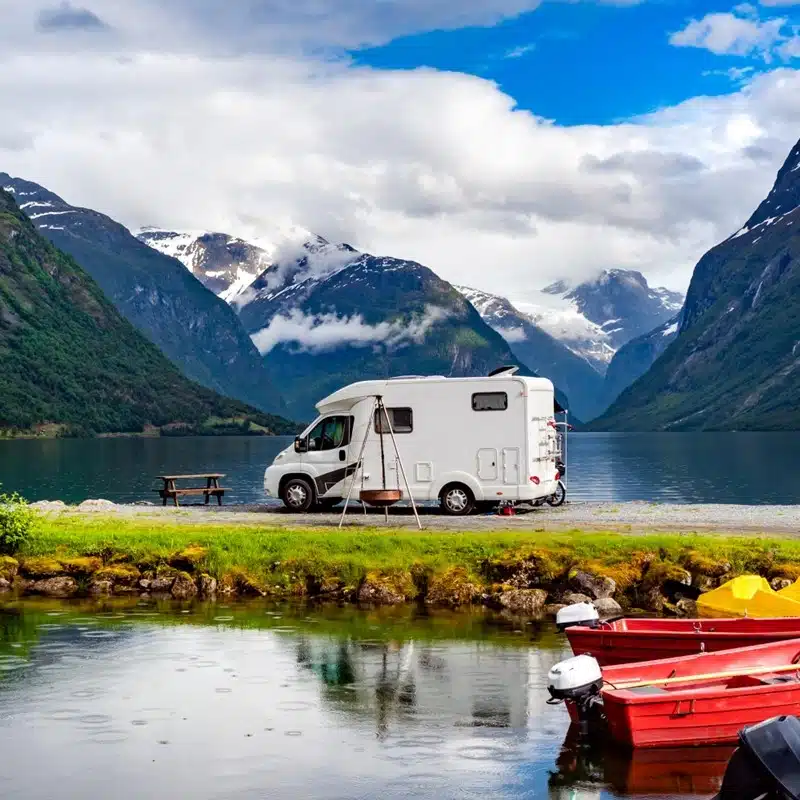 White RV parked by a scenic mountain lake with snow-capped peaks, small boats and a picnic table nearby