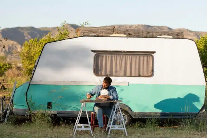 Man sitting at a folding table reading outside a vintage turquoise and white caravan parked in a dry rural landscape